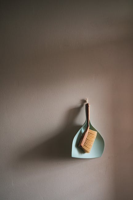 A muted grey wall featuring a minimalist hanging setup with a light blue, teardrop-shaped ceramic hook. Attached to the hook is a wooden-handled scrub brush with natural bristles, positioned neatly in