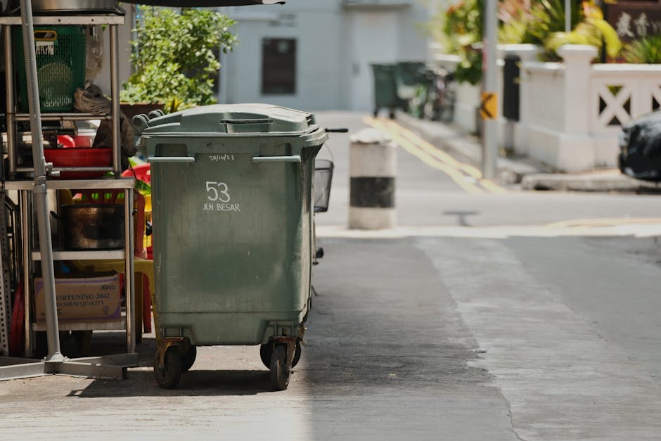 A worker wearing a white cap, green uniform, and a face mask is pushing a yellow wheeled bin for waste collection on a street with a black broom in hand. The bin has recycling symbols and Vietnamese t