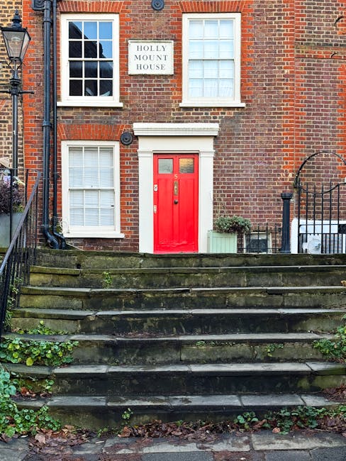 A narrow residential street in West Hampstead featuring a mix of brick and white-painted houses. The street is paved with cobblestones and asphalt, with a slight curve leading towards a cluster of tre