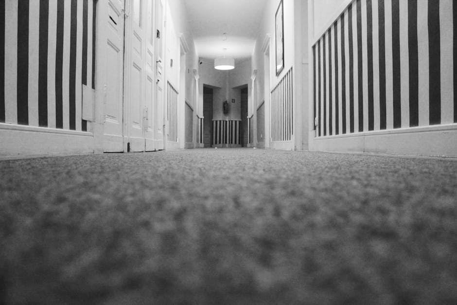 A professional cleaner from Cleaners West Hampstead is performing deep carpet cleaning in a residential living room. The cleaner is dressed in a white protective suit, gloves, a face mask, and shoe co